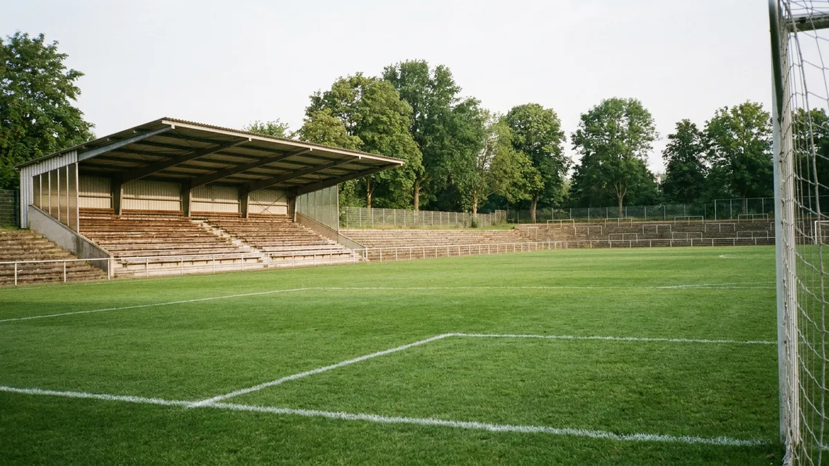 Leeres kleines Fußballstadion mit überdachter Tribüne und Stehstufen, rechts im Vordergrund ein Tornetz