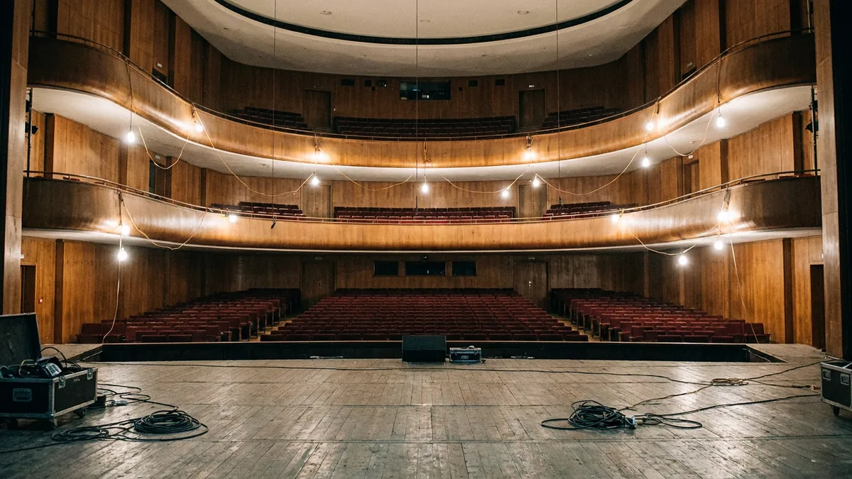 Blick von einer Bühne mit Kabeln und Kisten in einen leeren Zuschauersaal mit roten Sitzreihen und zwei Rängen