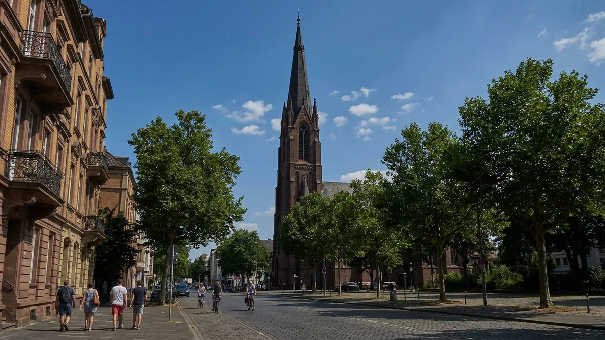 Neugotische Backsteinkirche mit hohem Einzelturm an einer baumgesäumten Kölner Straße im Sommer.